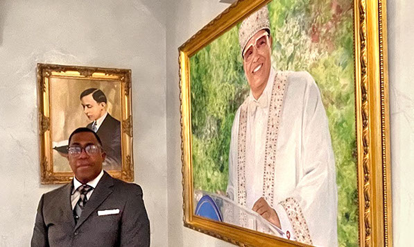 Carey Muhammad posing by Portraits of Master Fard Muhammad and the Hon. Minister Louis Farrakhan&nbsp; 